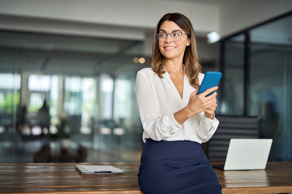 Smiling mid aged 40s professional businesswoman manager entrepreneur using cellphone working on smartphone.
